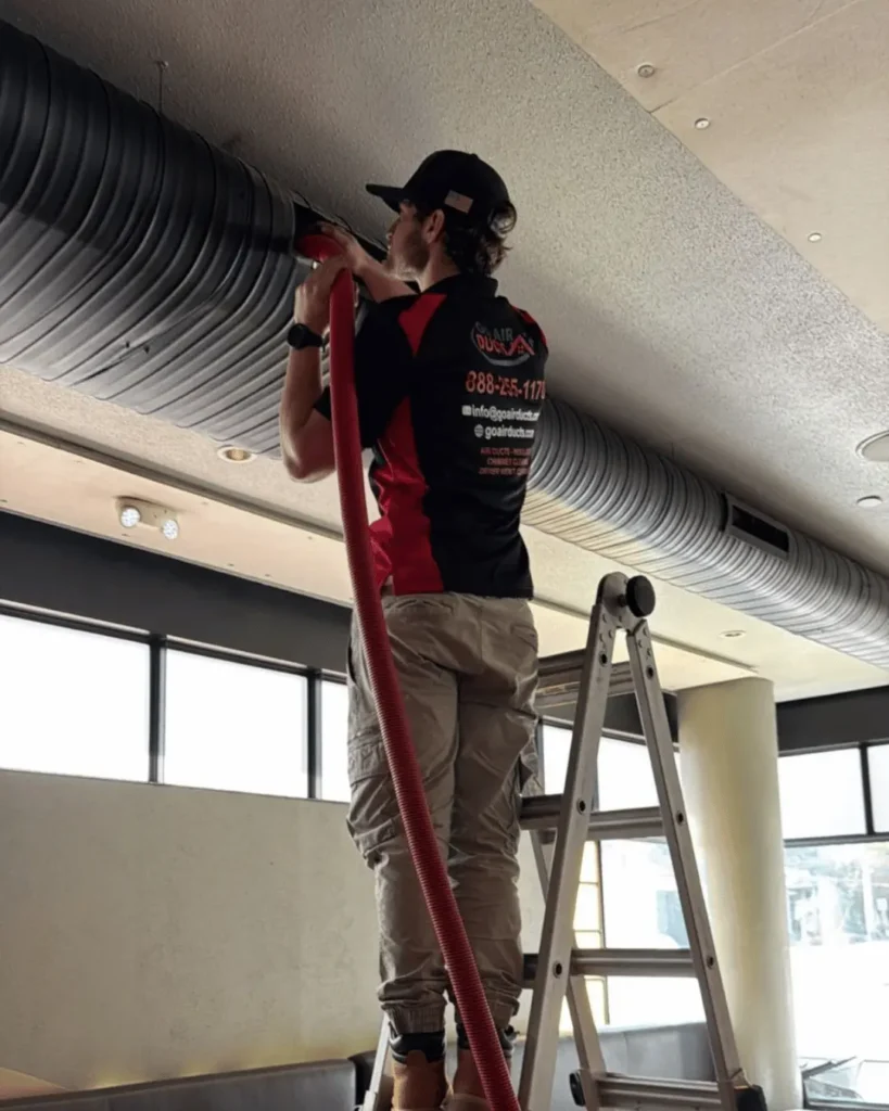 Technician cleaning air ducts with vacuum hose mounted on a ladder in a restaurant setting