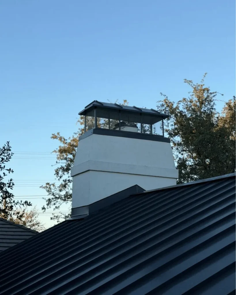 Chimney cap on a white residential chimney, surrounded by rooftops and clear sky