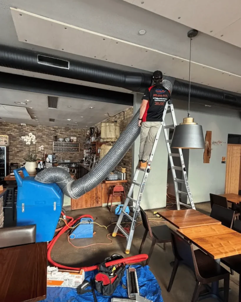 Technician cleaning air duct vent while on a ladder in a commercial dining area