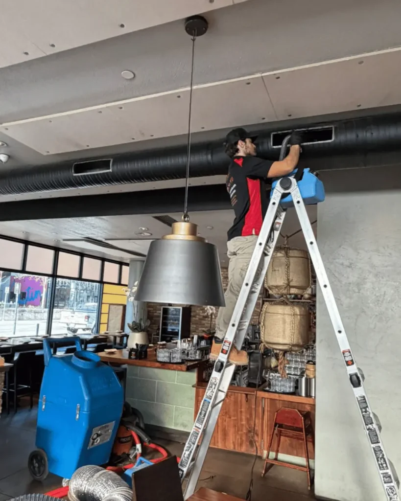 Technician cleaning commercial ductwork using an industrial vacuum system while on a ladder